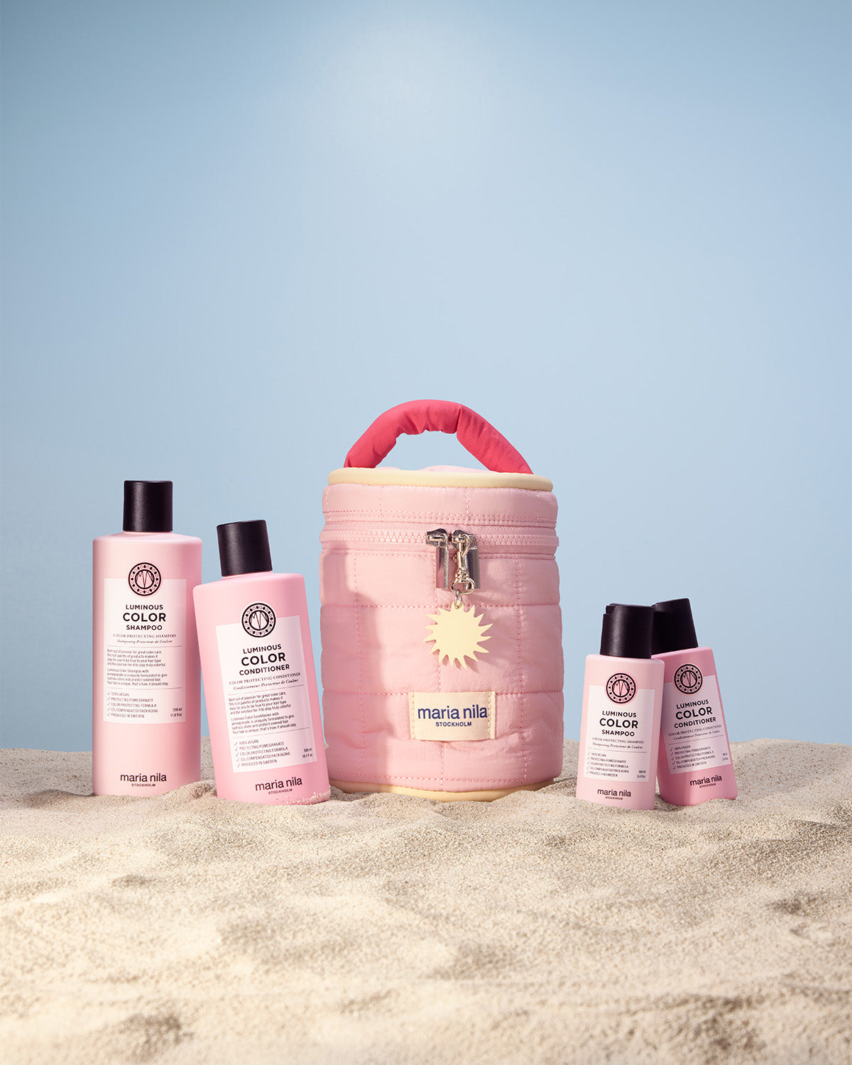 Set of Maria Nila hair care products including bottles and a Beauty Bag bag on sand with a blue sky background.