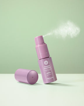 A lavender purple Maria Nila Power Powder bottle with its cap off, shown spraying with a visible mist cloud. The bottle is set against a clean, light green background on a matching surface.